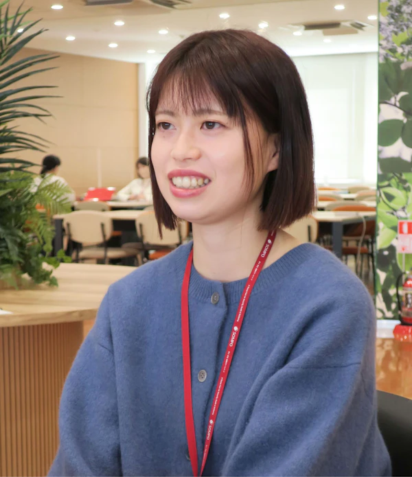 青いセーターを着た短髪の女性が笑顔で立っている。背景にはオフィスらしき明るい室内空間が見える。女性は赤いネックストラップの身分証を首から下げている。