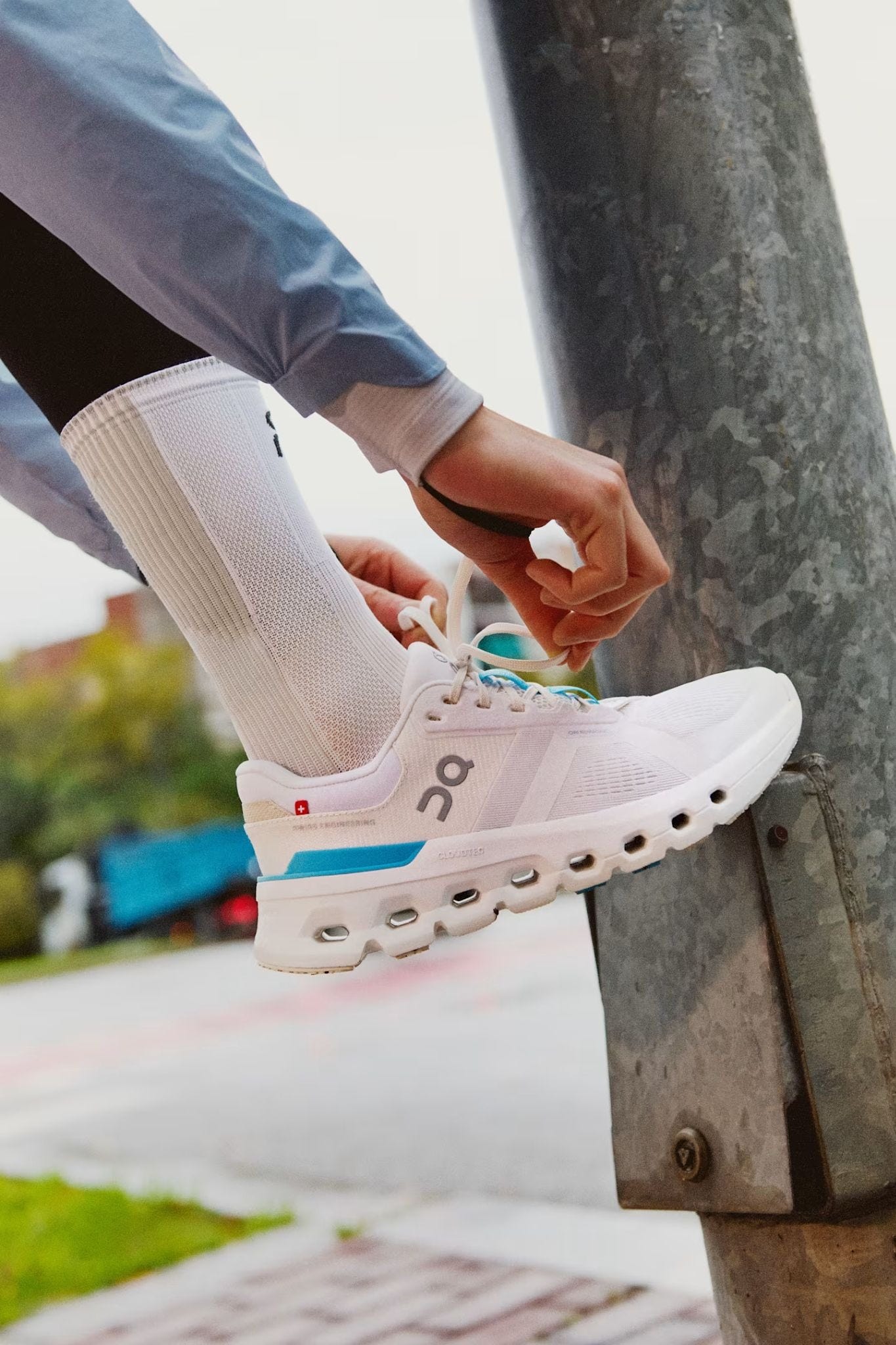 Onの白いランニングシューズを履いた人が街角の支柱に足を乗せ、CloudTecソールが見える状態で靴ひもを結んでいるクローズアップ写真。