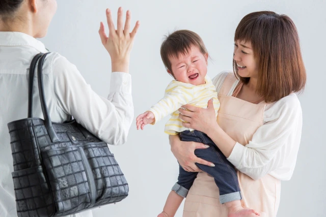 泣く子供を抱っこする保育士さんと手を振るお母さん