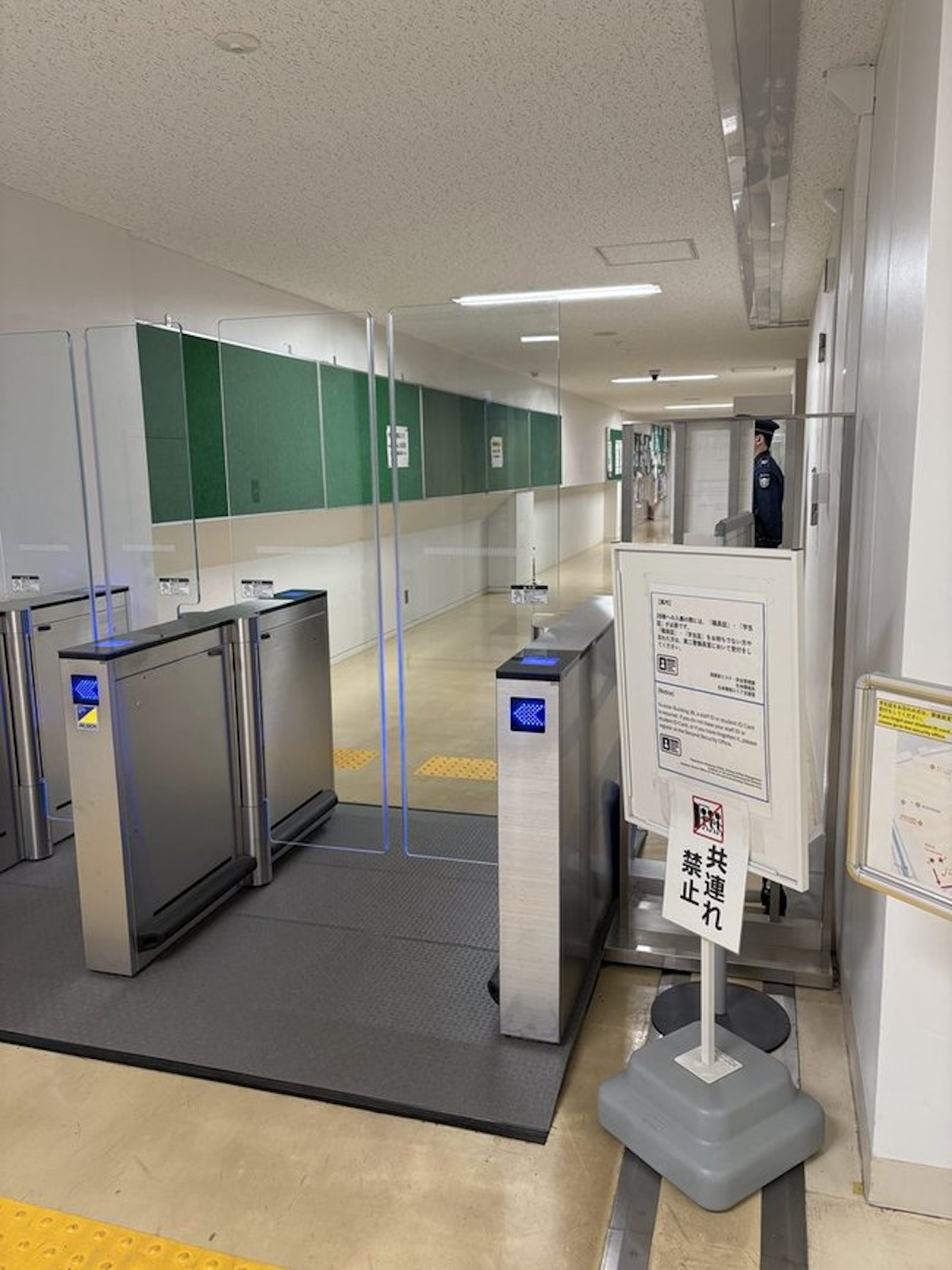 筑波大2B棟に設置された警備用ゲートの写真。外から中の方を撮影している。奥には警備員の姿も見える。ゲートの横には「共連れ禁止」などと書かれた注意書きの看板がある。