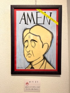 壁にかけられたイラスト作品の写真。「AMEN」「MAN of the YES?」の文字と日本の首相に似た男性が描かれている。