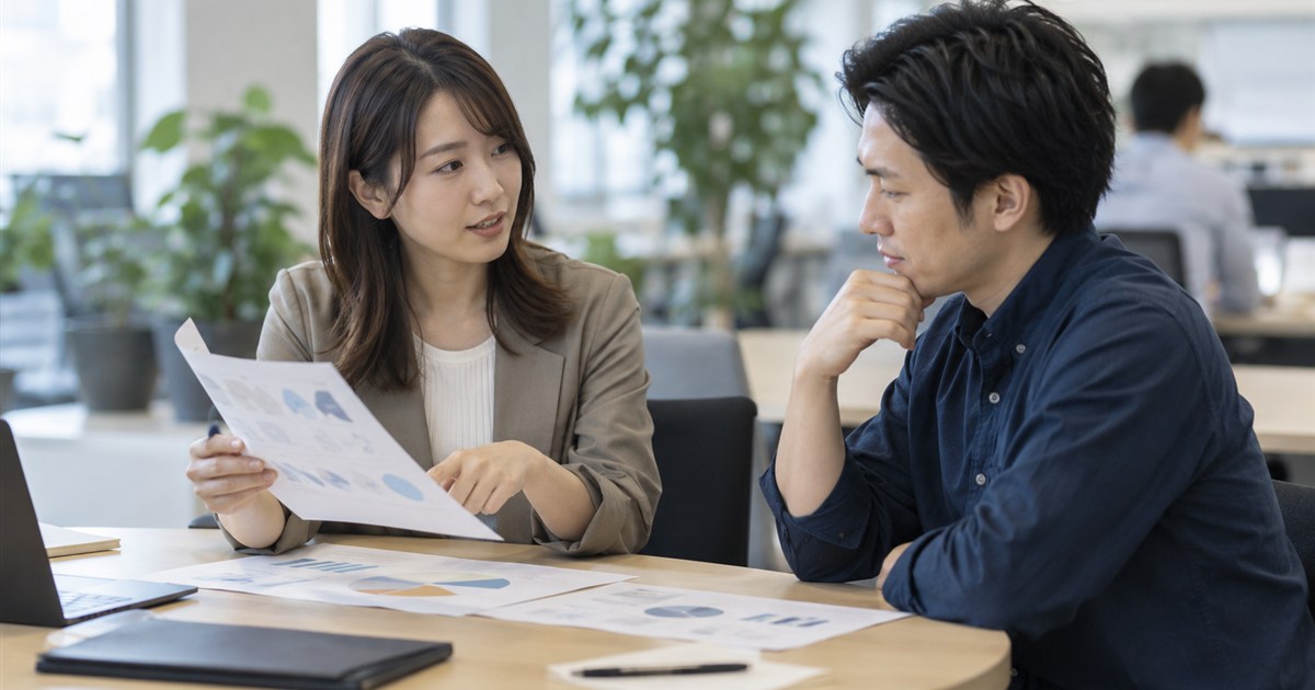 社内の打ち合わせスペースで、マーケティング担当者が技術部門の社員に資料を見せて相談している様子