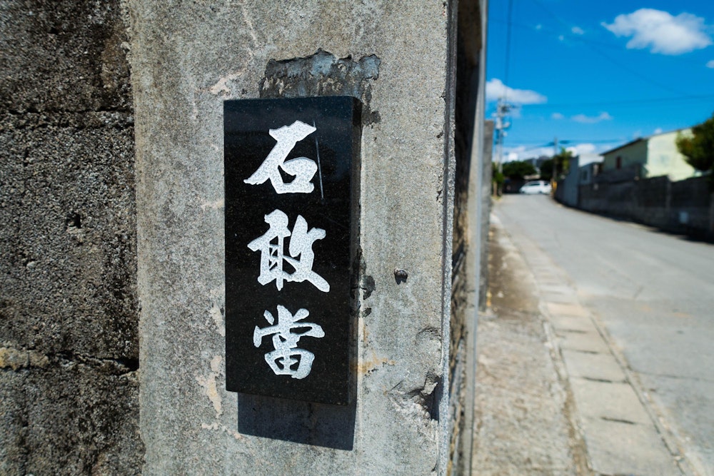 <p>「石敢當」。無知な自分は、これを見て珍しい苗字の人がいるものだなと最初は考えた。<br>しかし、歩き進めているとそこらじゅうで見かけることになり、これは表札ではなく、なにか現地の文化的なものだと悟る。<br>調べてみると、これはマムジンという古くから沖縄で言い伝えられている魔物を除けるためのお札のようなものだという。<br>真っ直ぐにしか進めないマムジンは壁にぶつかるとそのまま家の中に入ってきてしまうため、道路が突き当たる箇所にこの石敢當を配置することで、それを防ぐのだそう。</p>