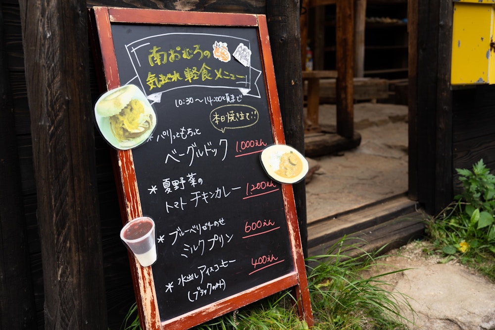 <p>南御室小屋に到着すると目に入ってきたのは小屋の前に出ていた軽食メニュー。<br>どれも目を惹かれたが、ベーグルドックとブルーベリーのせミルクプリンをいただく。<br>2 人で同じものを食べて一緒に感動。どちらも絶品で、ここまでの行程を歩いた疲労が吹き飛ぶ。<br>今日はここで幕営したが、テント場もこじんまりとしていて落ち着く場所だった。</p>