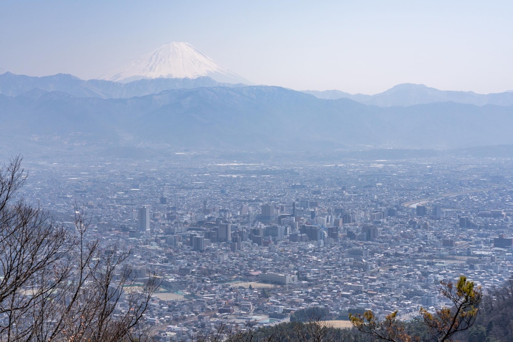 <p>甲府市中心部のビル街の向こうに霞む御坂山地、そしてその奥にうっすらと見える雪を被った富士山。<br>甲府から見る富士山は御坂山地に遮られているものの、やはり距離が近いだけあって大きく見える。</p>