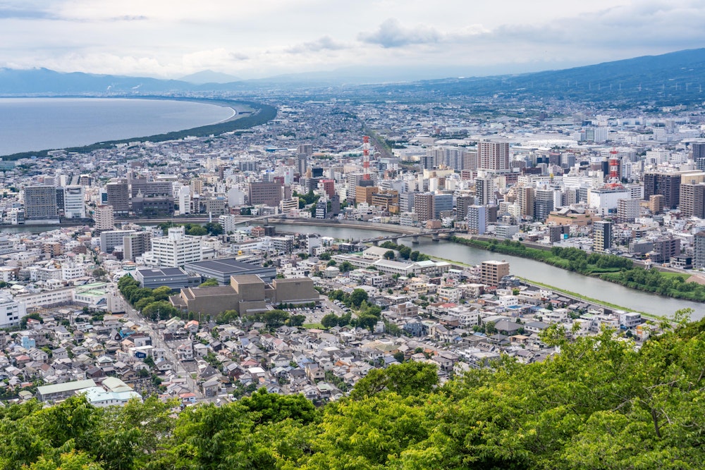 <p>沼津市中心部方面。手前を流れる川は狩野川。<br>こう見ると沼津市は意外とマンション化率が高い都市に見える。<br>左奥は駿河湾、右奥のゆるやかな斜面は富士山の裾野。<br>この日はあいにく雲が多く富士山は顔を出していなかった。</p>
