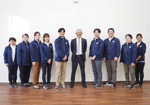 Shuichi, in a suit, stands in the middle of the picture with nine staff members standing on both sides of him.