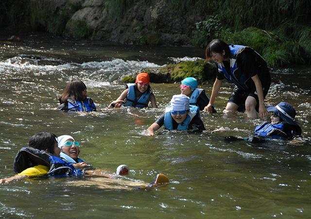 Children wearing life jackets are playing in the river with staff members.