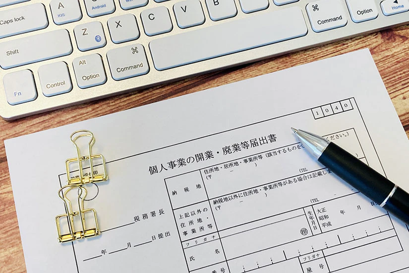 木目の机の上に置かれた個人事業の開業届とペン、キーボードの画像