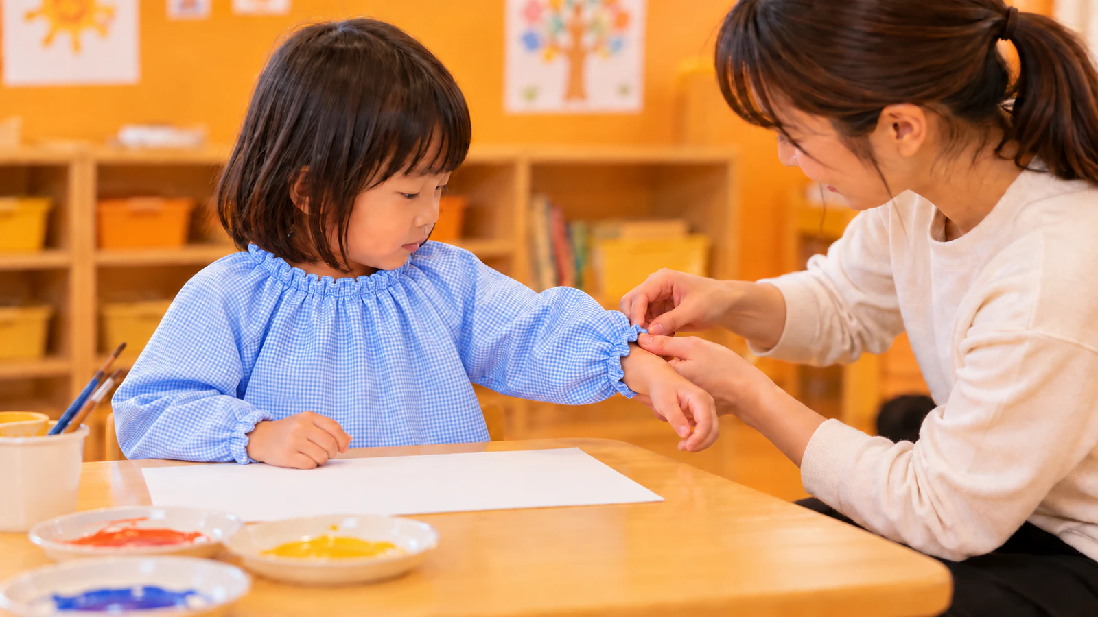 保育室の机の前で、水色のギンガムチェックのスモックを着た子どもの袖口を、先生が手首に合わせて整えている様子