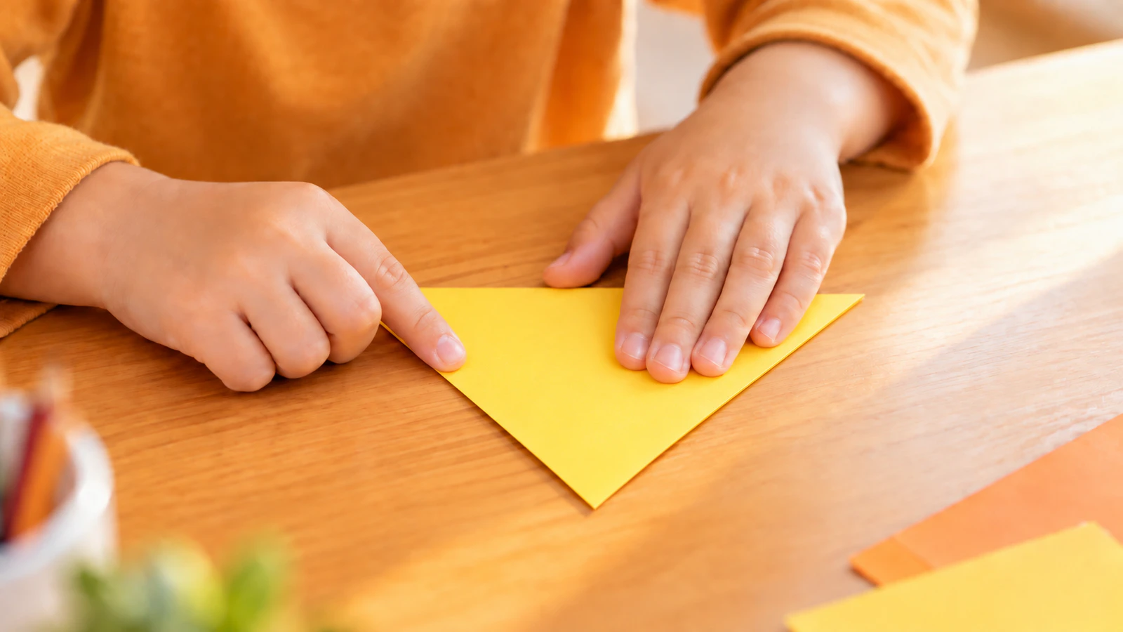 子どもの手が黄色い正方形の紙の角と角を合わせ、三角に折って指で折り目を押さえている手元