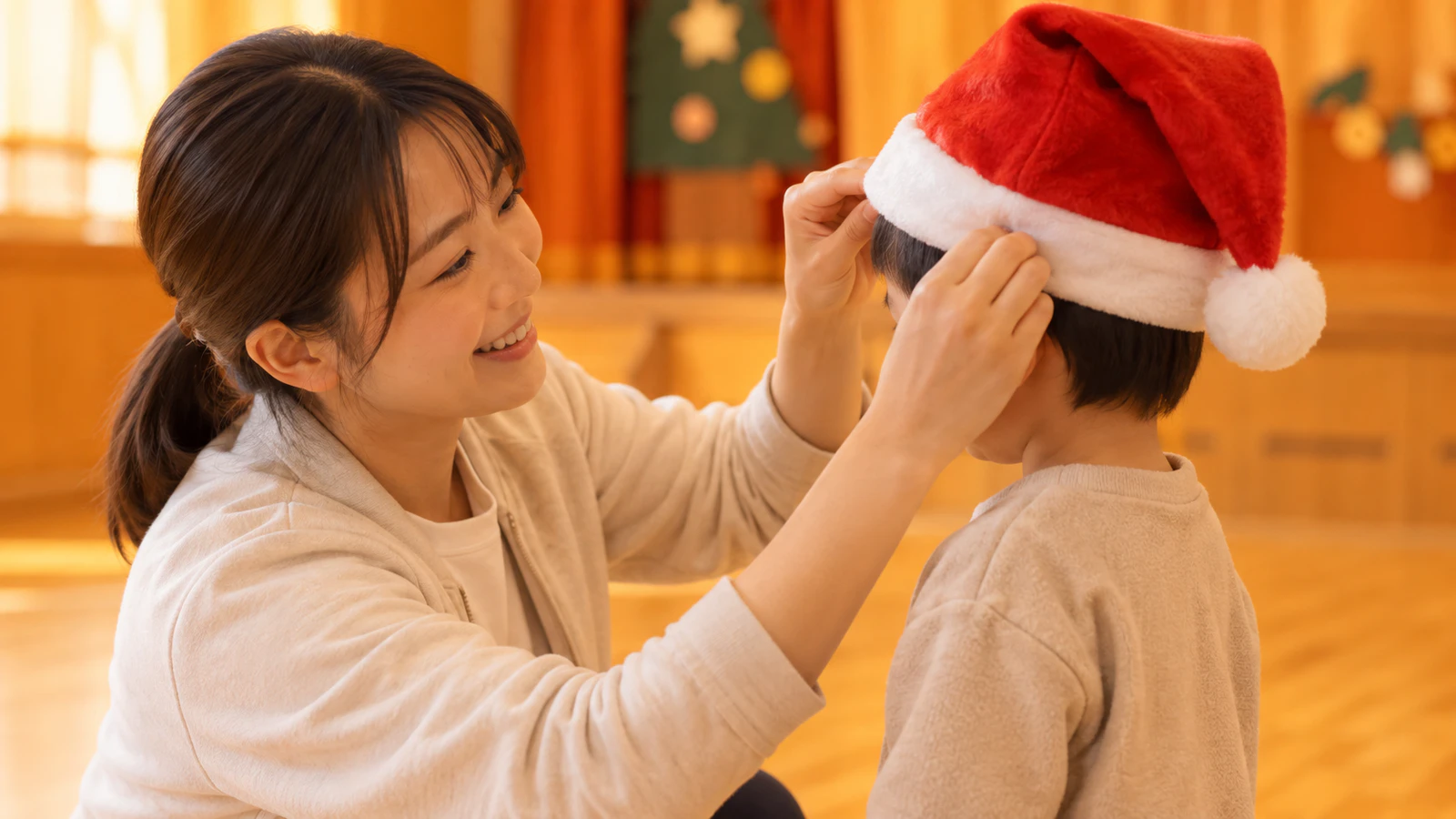 園のホールで、先生が普段着の子どものかぶったサンタクロースの帽子の位置を整えている様子