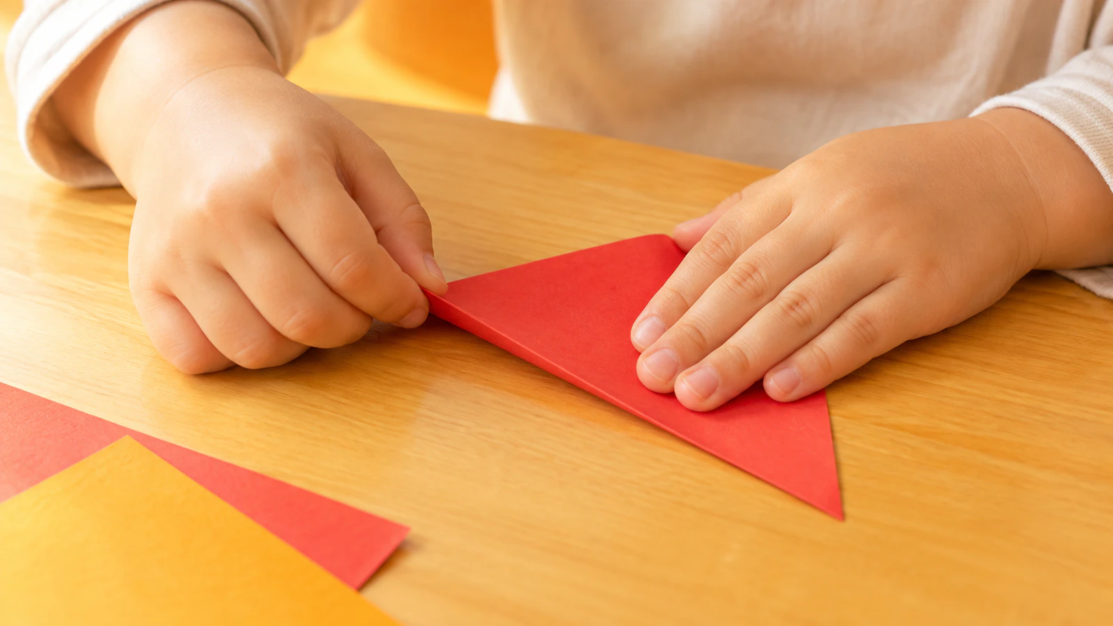 子どもの小さな手が、赤い折り紙の角と角を合わせて折り目をおさえている手元