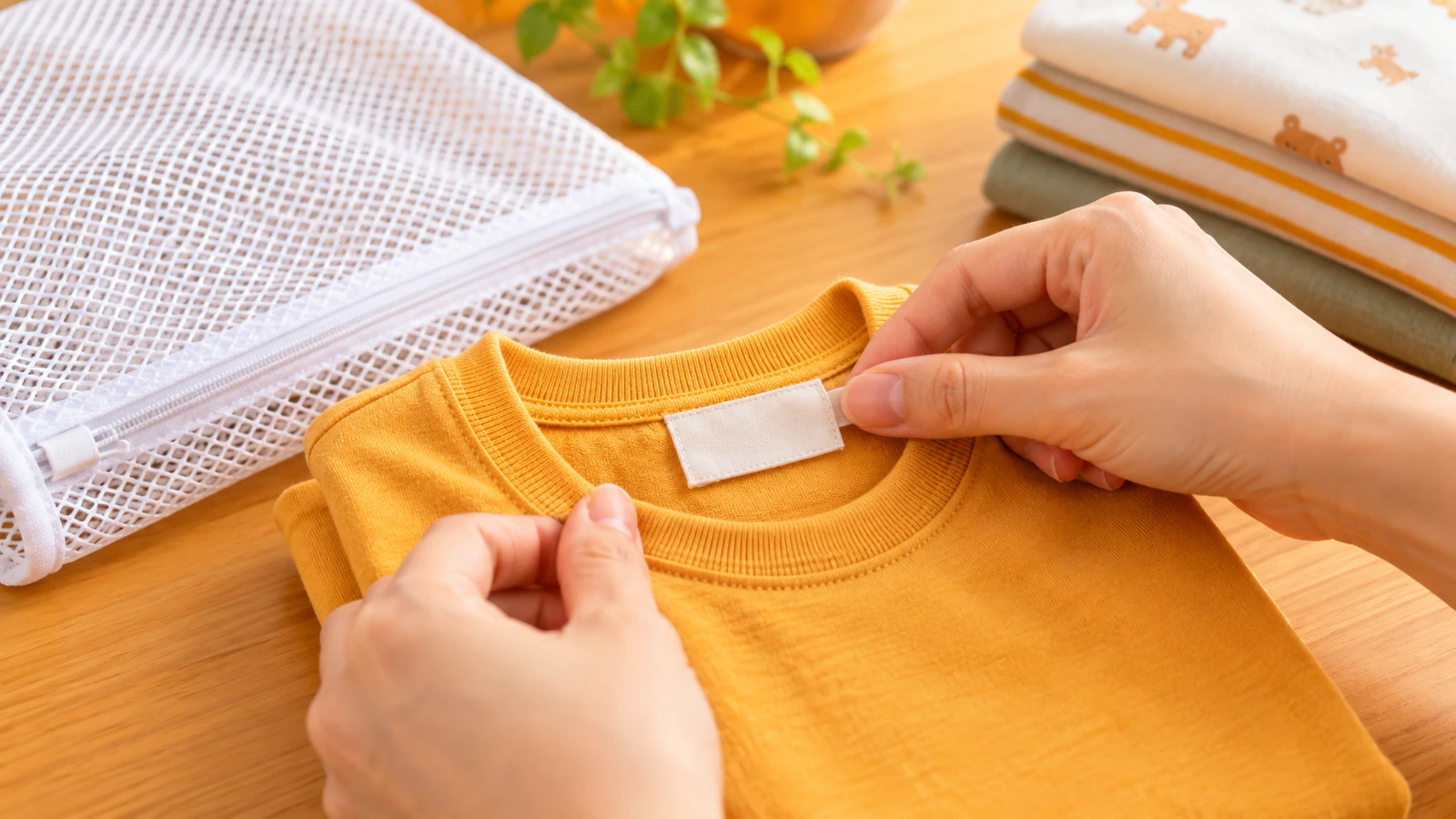 洗濯して畳んだ子ども服の首の後ろにある無地の名前布を、大人の指で確かめる手元と白い洗濯ネット