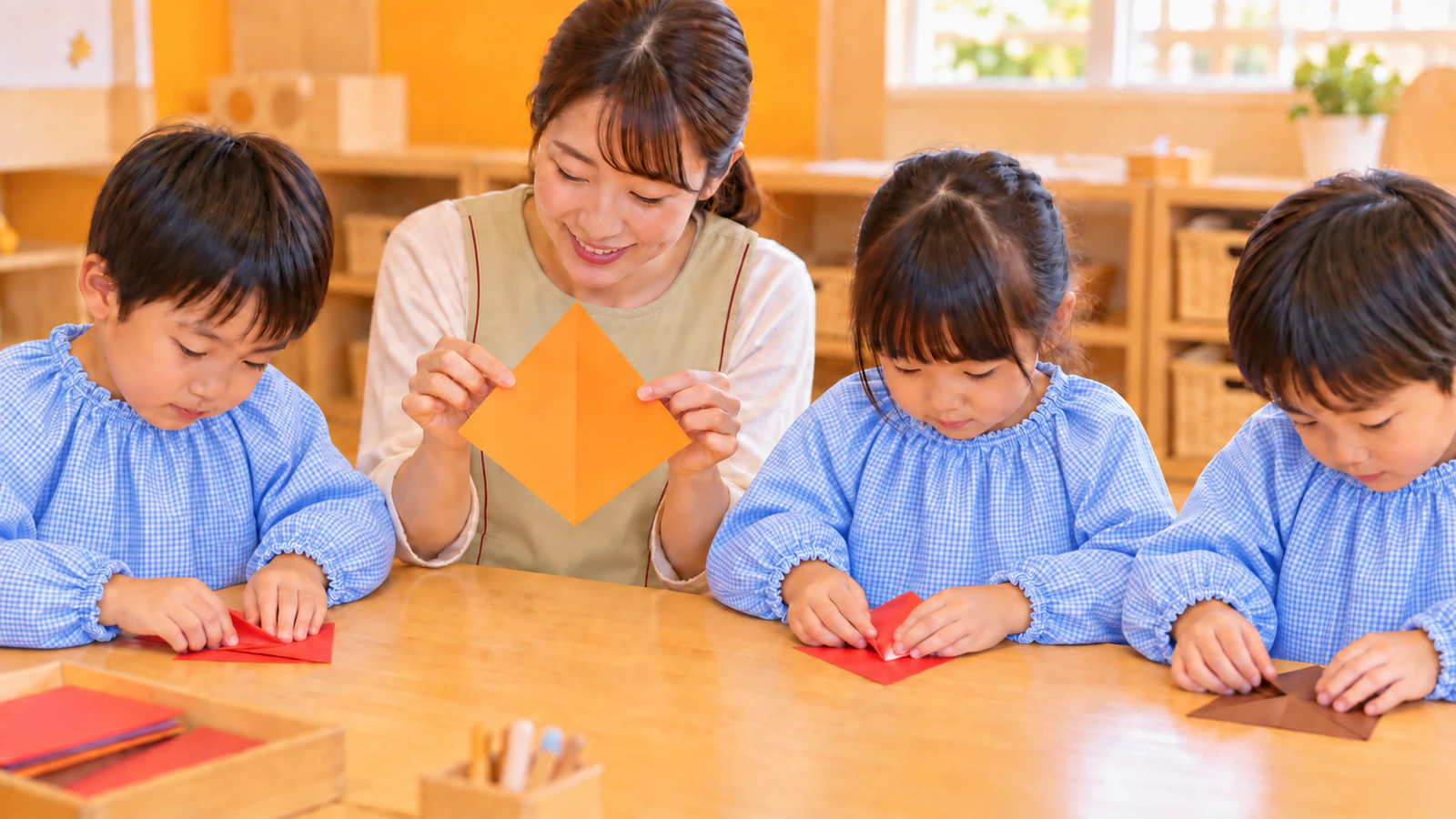 保育室の低い机で、子どもたちと同じ側に並んで座った先生が大きなオレンジの紙をひし形の向きに持って見せ、スモックを着た子どもたちが自分の赤や茶色の折り紙を折っている様子