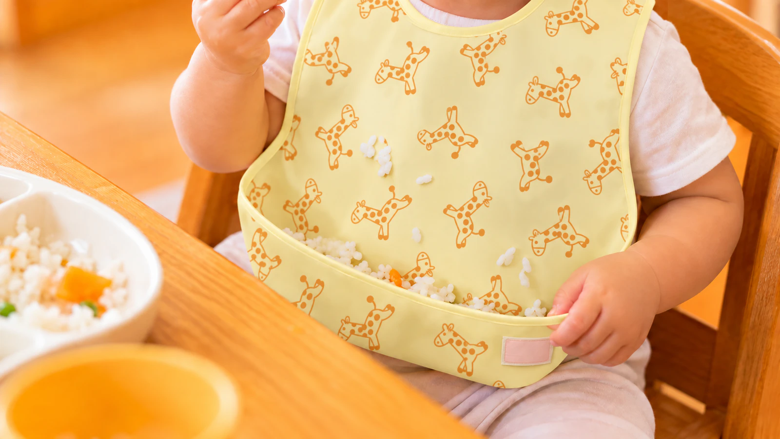 テーブルに座る子どもの胸元の寄りで、キリン柄の袖なしエプロンのポケットが、こぼれたご飯粒を受け止めている様子