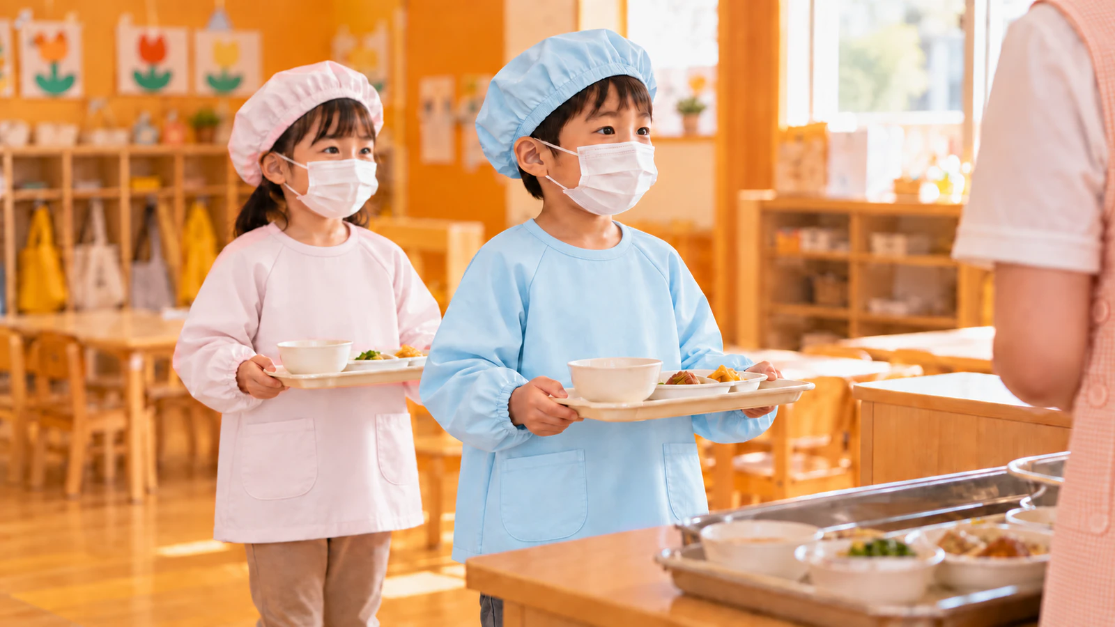 保育室で、水色と薄いピンクの給食着に帽子とマスクを着けた5歳ごろの子どもたちが、おぼんを持って配膳台に並ぶ様子
