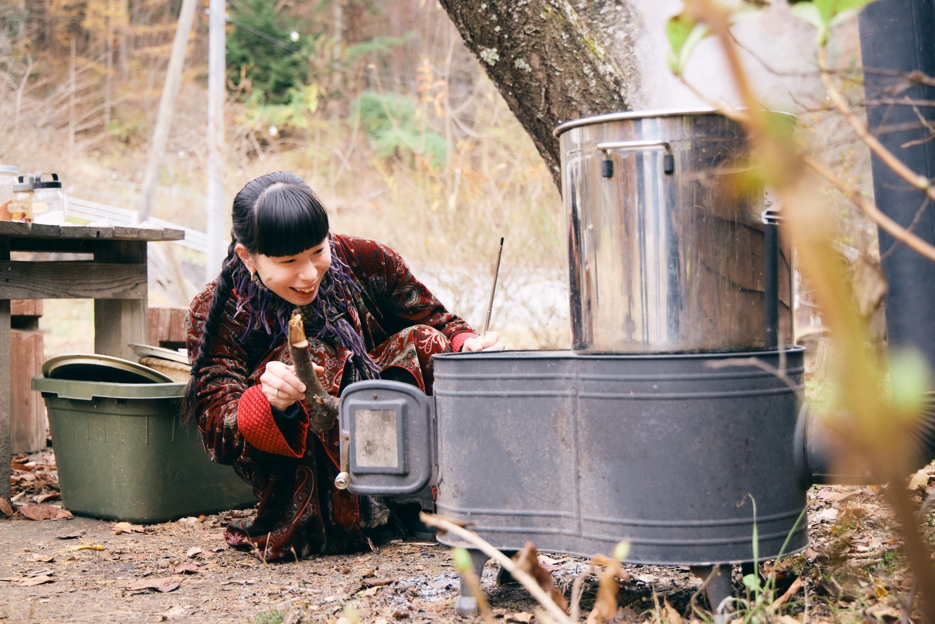 A photo of Ms. Ushiozu opening the door of a wood-burning stove that is heating a silver pot and burning a roll.