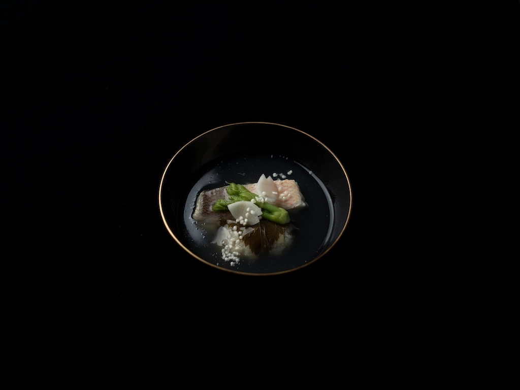 A soup consisting of tilefish and fatsia sprouts in a black lacquered vessel featuring a gold rim.