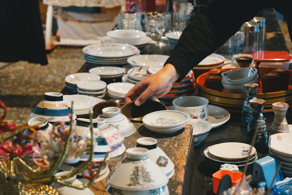 A photo of a man trying to pick up a plate lined up in a store. The store is lined with countless numbers of teacups, plates, and tokkuri (Japanese rice wine cups).