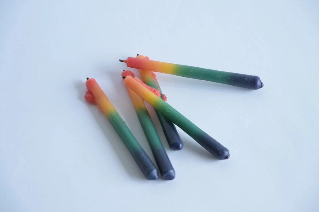 Photograph of five rainbow-colored candles. Wax is dripping from the tips of several of them and hardening.