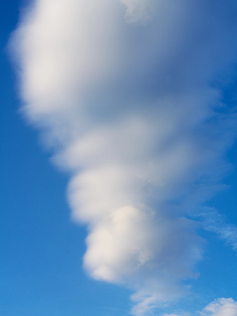 Photo of white clouds floating in a clear blue sky. The clouds extend vertically, narrow at the bottom and gradually grow wider further up the photo, diffusing as they widen.