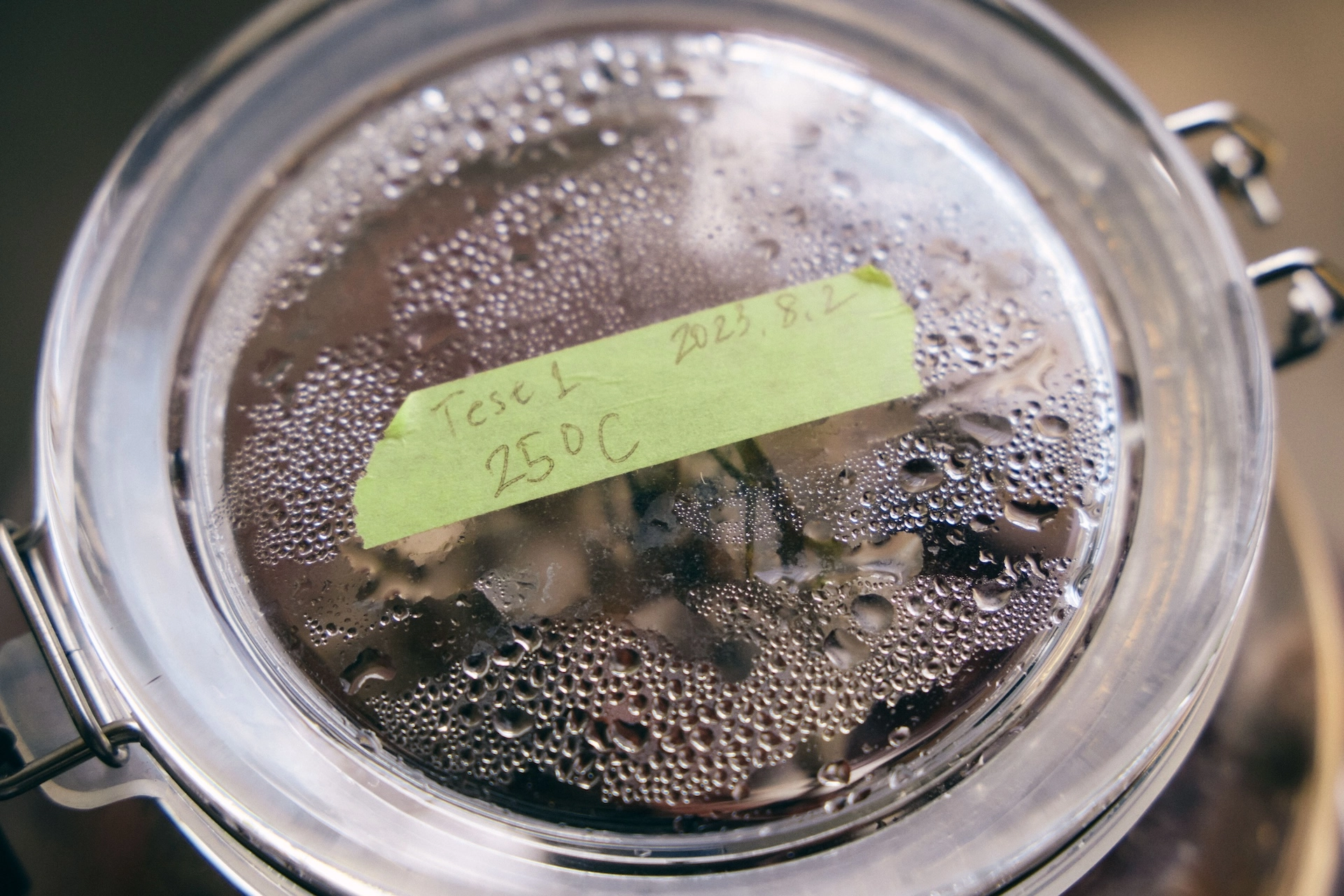 Green tape is affixed to the lid of the glass vessel. The handwritten text reads “Test 1 25°C 2023.8.2.” There are water droplets on the back of the lid.