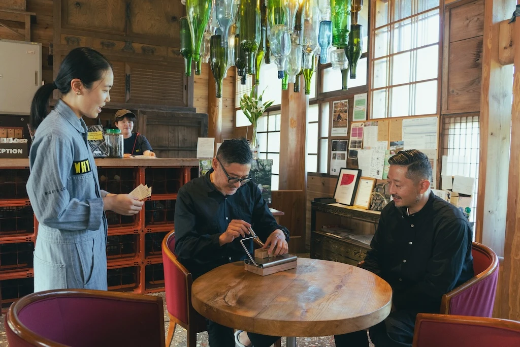 固形石鹸をスライスしている色部さんとそれを横で見ている渡辺さん。大塚さんだけ立っており2人に何か説明をしている写真。