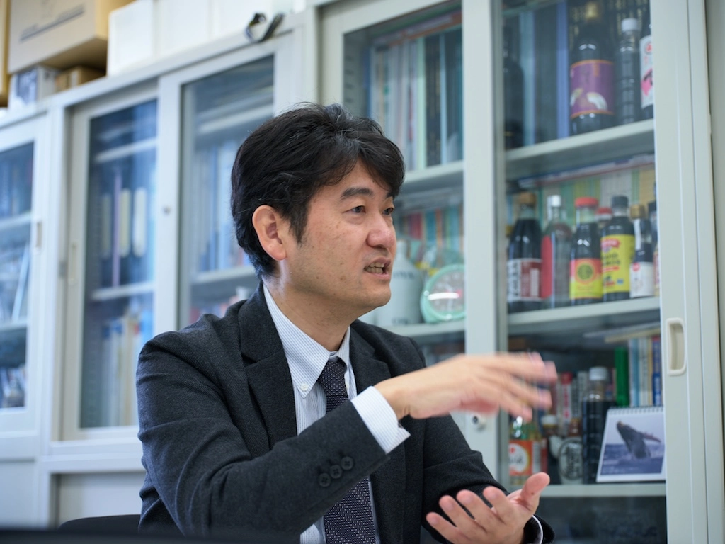 Professor Maehashi speaks while sitting in a lab room.
