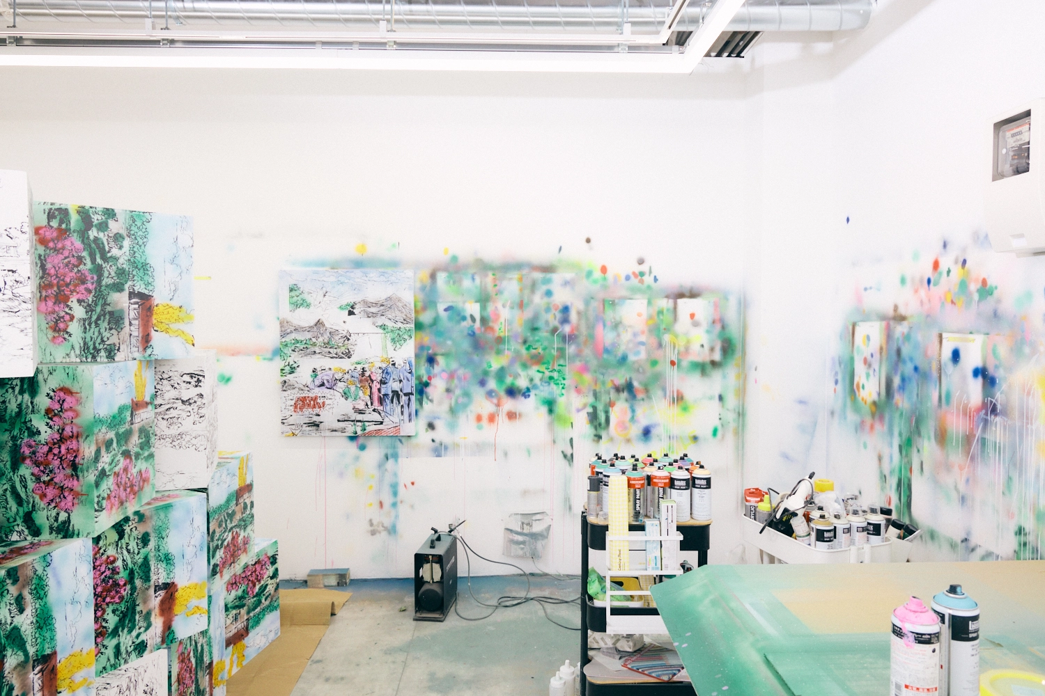 A photo of the studio. The walls are covered with sprays of various colors, and the carts are filled with sprays used for the artwork. Part of the artwork is visible on the left.