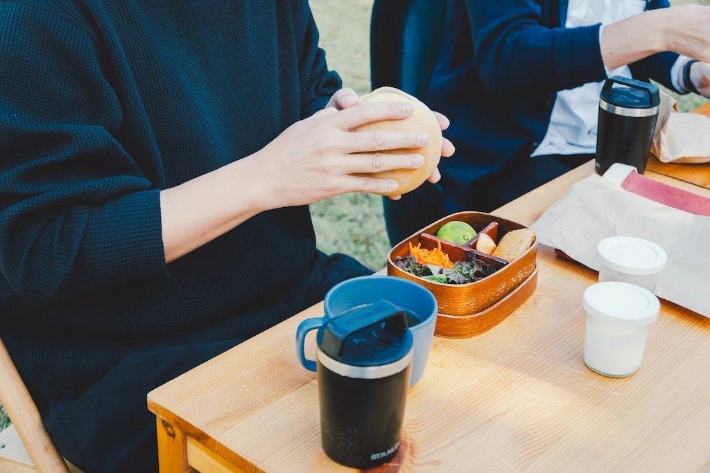 A photograph of a hand eating lunch outside. A lunch box, water bottle, and cup are on the table, and the figure is holding a loaf of bread.