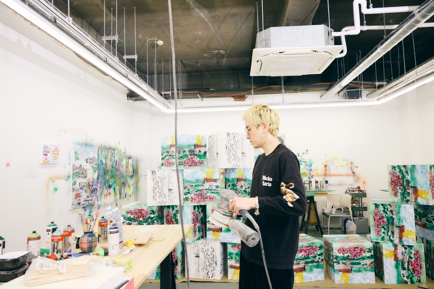 A photo of Hiroki Niimi working in his studio. He is standing in front of a worktable, holding a sander. His artwork is displayed in the background.