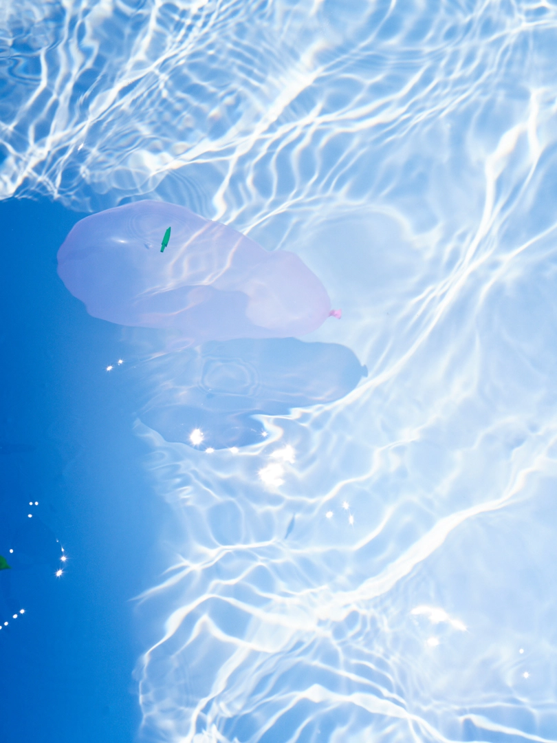 Photo of a light pink water balloon floating in water. The surface of the water sparkles with reflecting light, while ripples of shadow and light trace the bottom of the water.