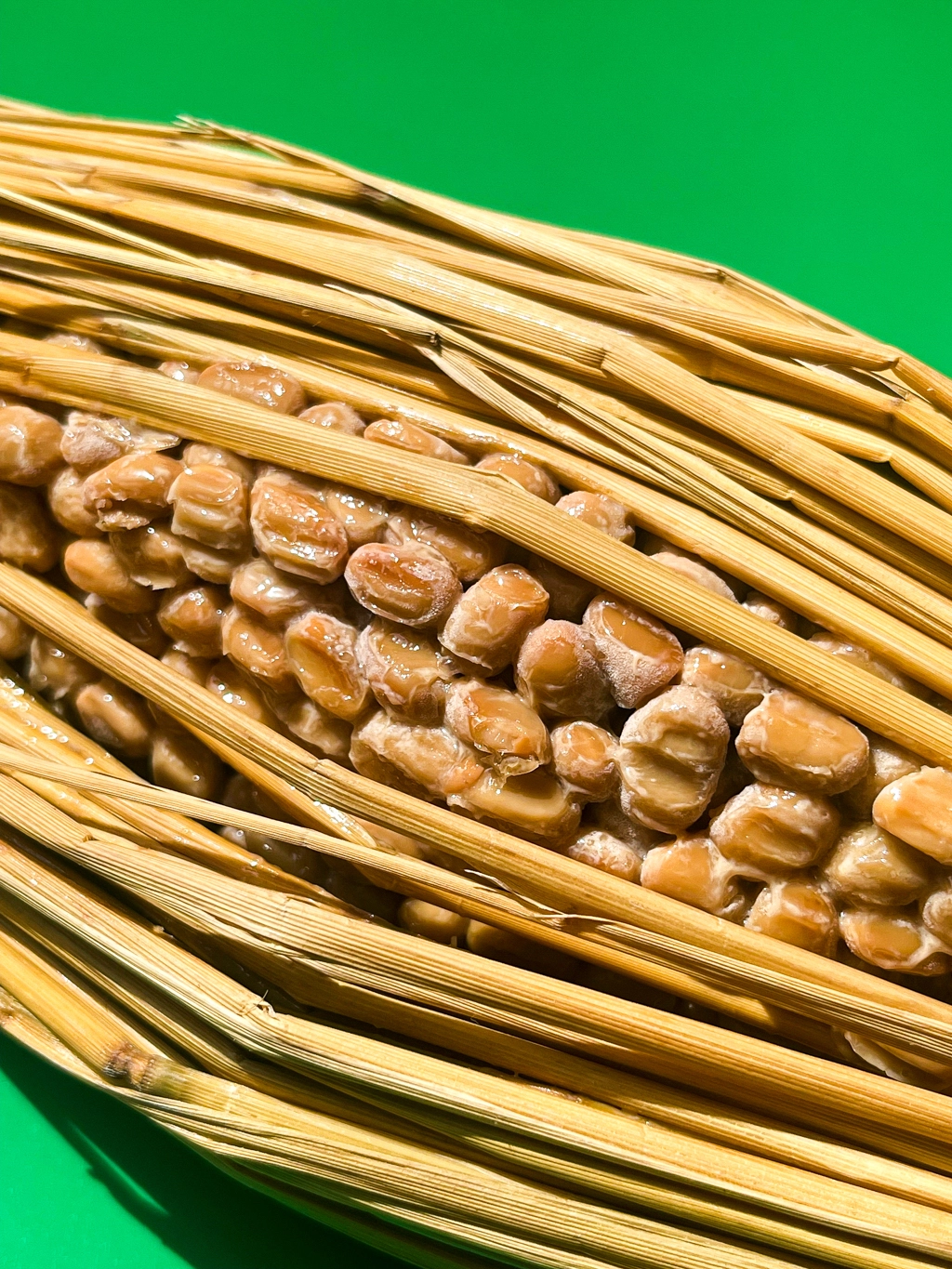 Artwork showing a close-up of natto wrapped in straw. Natto beans are visible through the slightly open straw. The background is a brilliant green.
