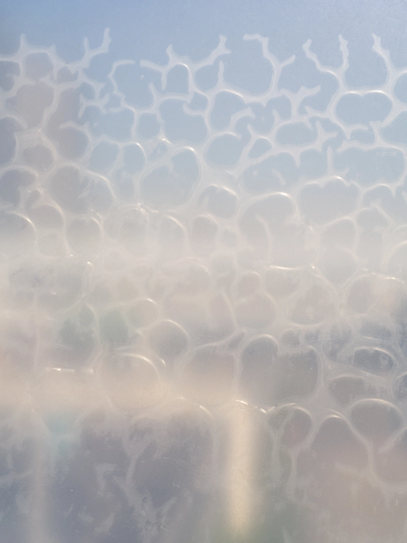 Photo showing a mesh-like pattern formed by a distorted film adhered to a glass-like object. An irregularly shaped cloudy white pattern appears on a transparent background. The pattern is random, and subtle colors slip in and out of view due to the refracting light.