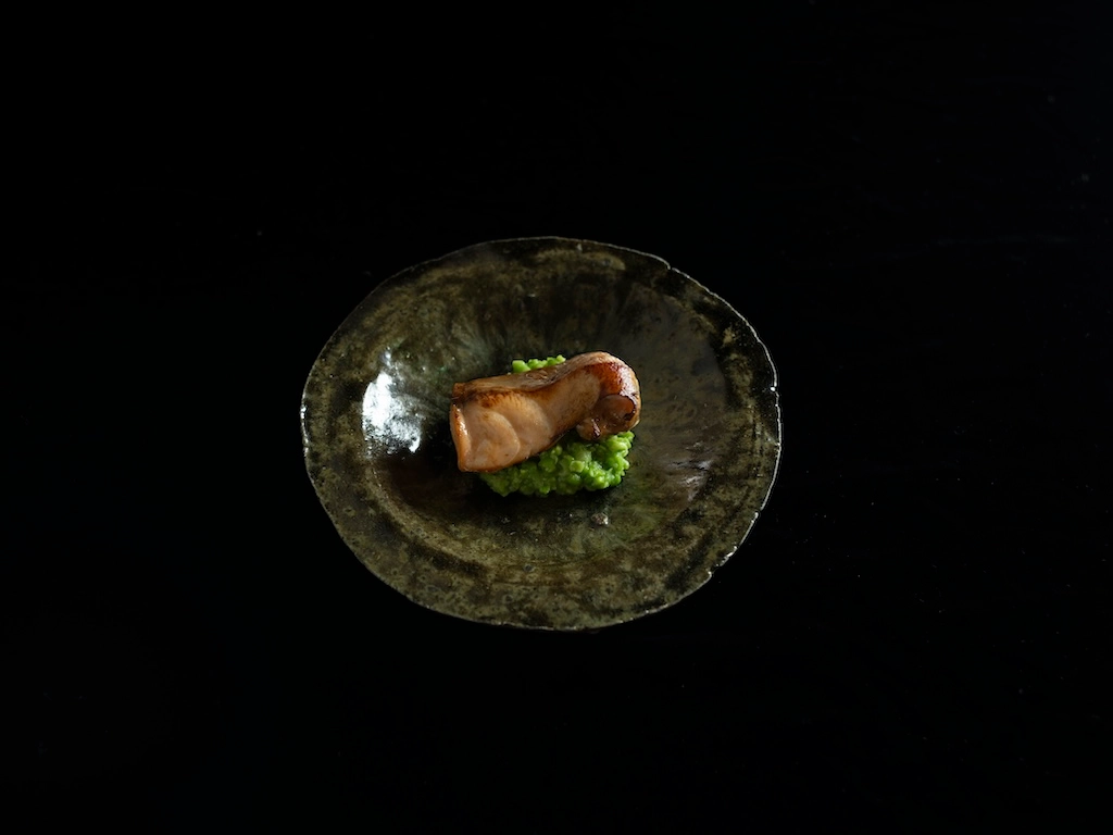 Grilled wild cherry trout served atop bright green pea paste, arranged on a circular vessel featuring a dark, uneven glaze with a slightly green hue.