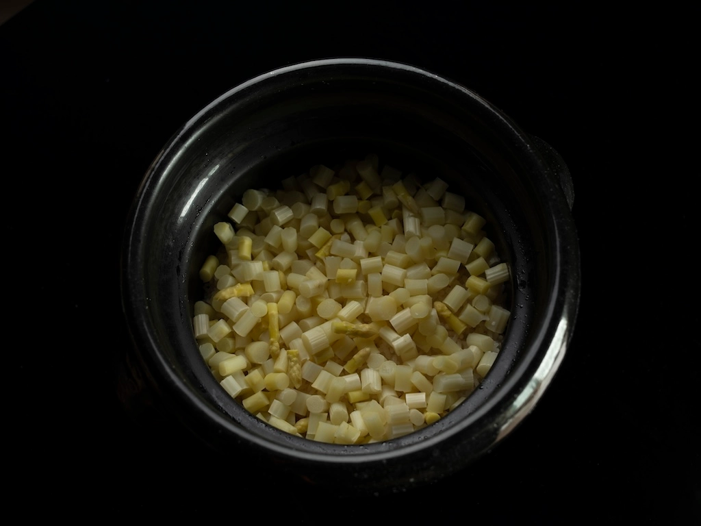 White asparagus rice cooked in a black clay pot. The chopped asparagus is laid out in the pot so as to cover the rice completely.