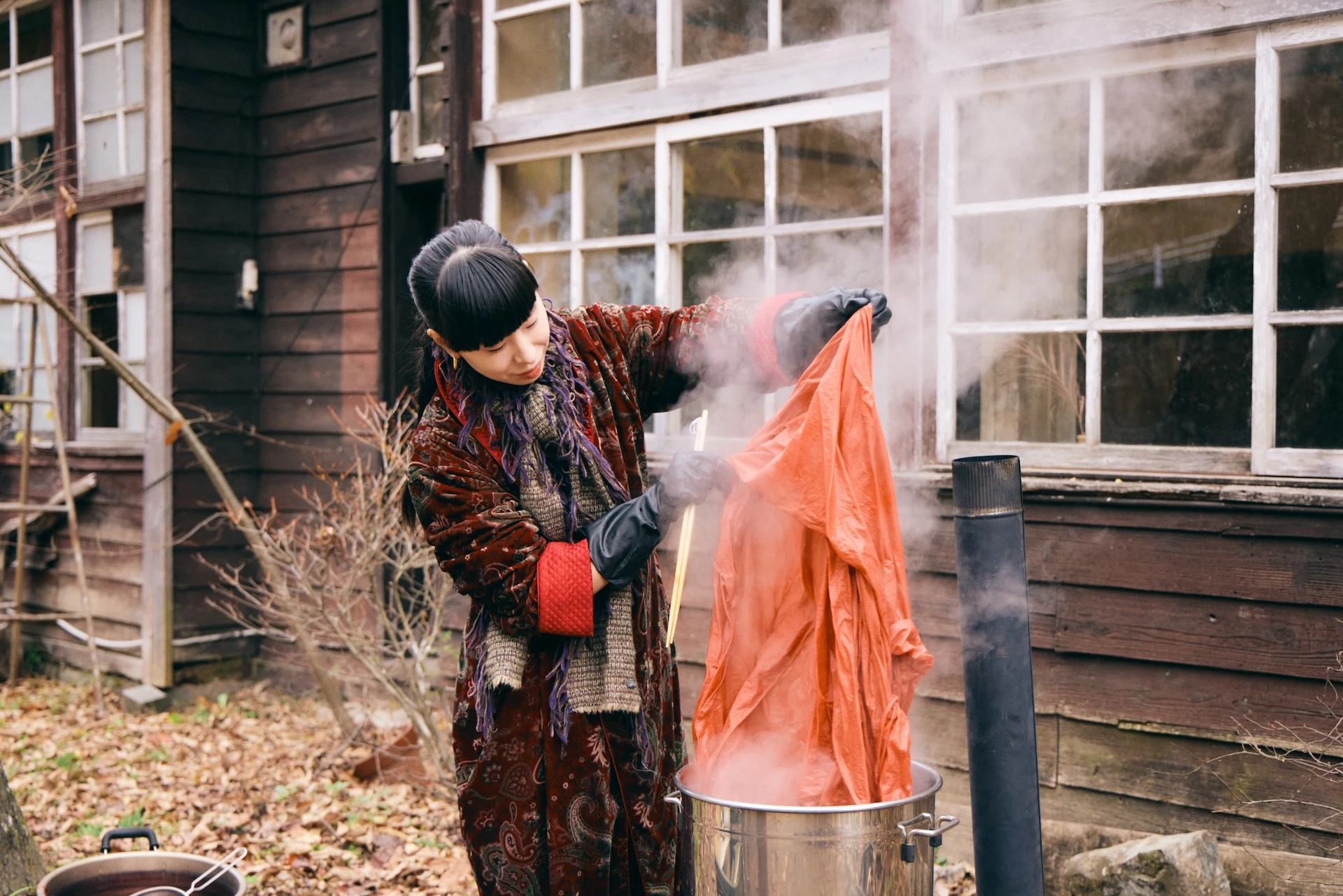 A photograph of Ms. Ushiozu as she removes a vermilion cloth from a silver pot. Steam is coming out of the pot and Ms. Ushiozu has rubber gloves on both hands.