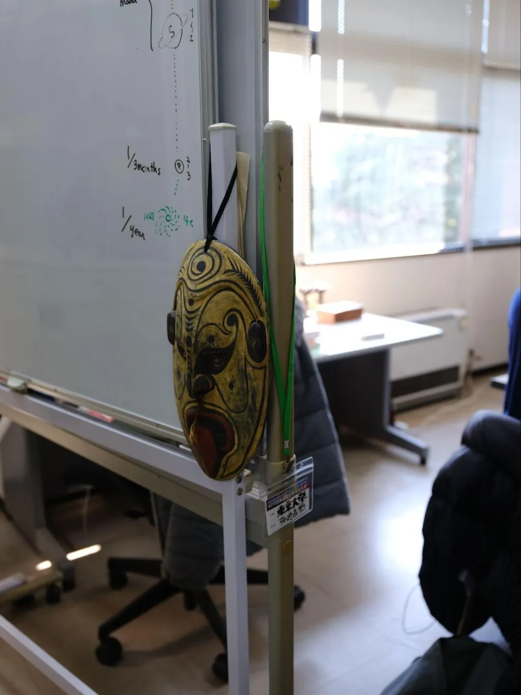 Photo of a yellow mask from an ethnic group in Papua New Guinea hanging on a whiteboard support in the lab. The mask has a long elliptical shape vertically and is covered in patterns. The mouth is red and shaped like it is sticking out its tongue.