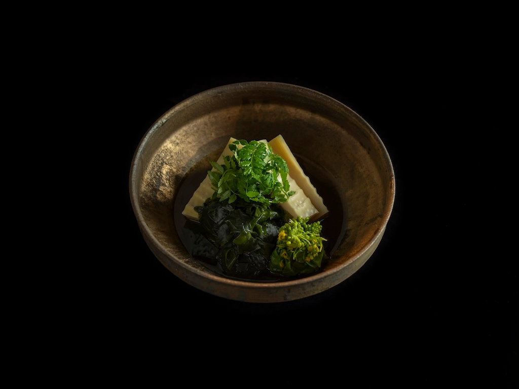 Boiled wakame seaweed and bamboo shoots served in a round brown vessel. This dish is also garnished with rapeseed blossoms and Japanese pepper leaves.