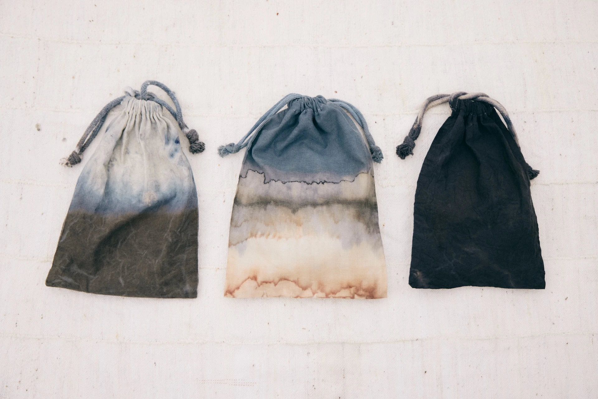 Photographs of three drawstring bags with different patterns dyed by herb dyeing. The left pouch has a light, almost white top with gradations of bluish gray and dark brown toward the bottom. The middle pouch is a dull blue at the top with cream and brown speckles toward the bottom. The right pouch is almost entirely dark black with discreet unevenness.