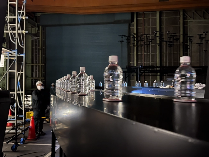 ベルトコンベアの上のペットボトルの群れを見守る関係者一同の写真