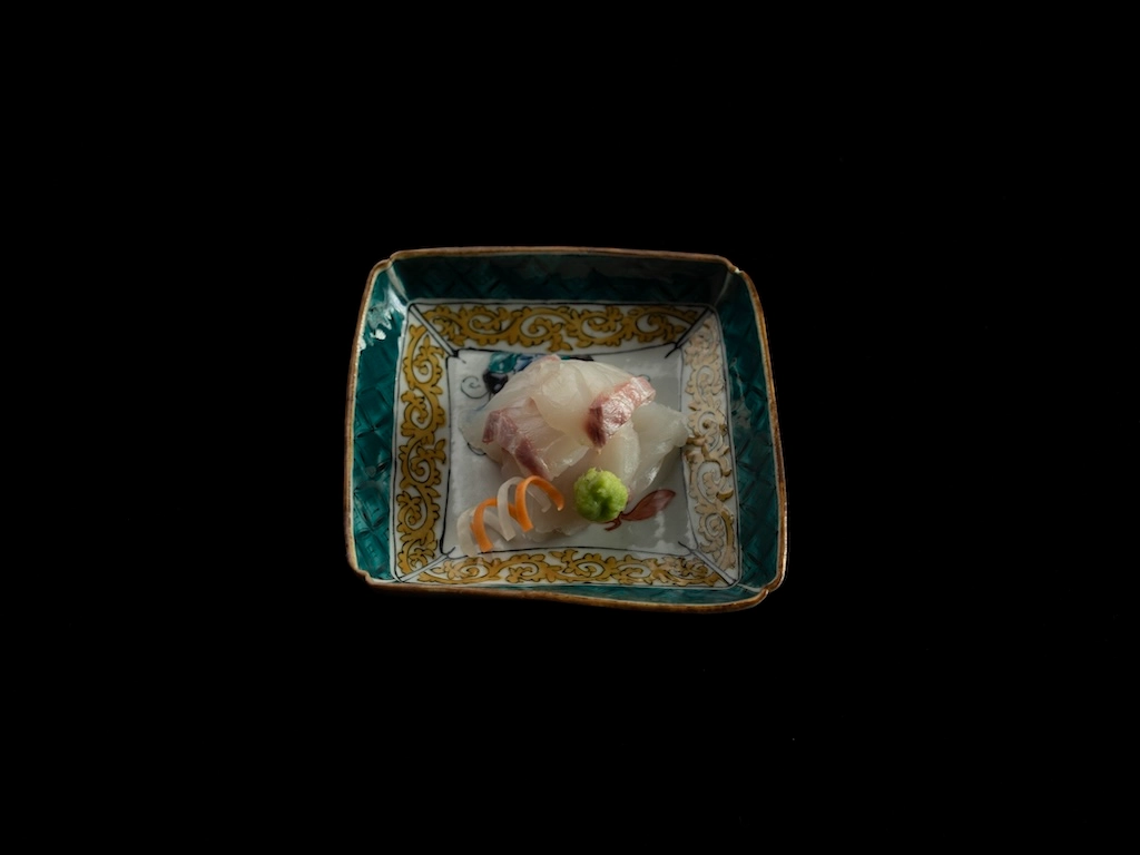 Red seabream sashimi served on a square vessel. It is garnished with wasabi and spiralized carrot and daikon radish. The ceramic vessel is painted with a design reminiscent of Japanese or Chinese motifs.