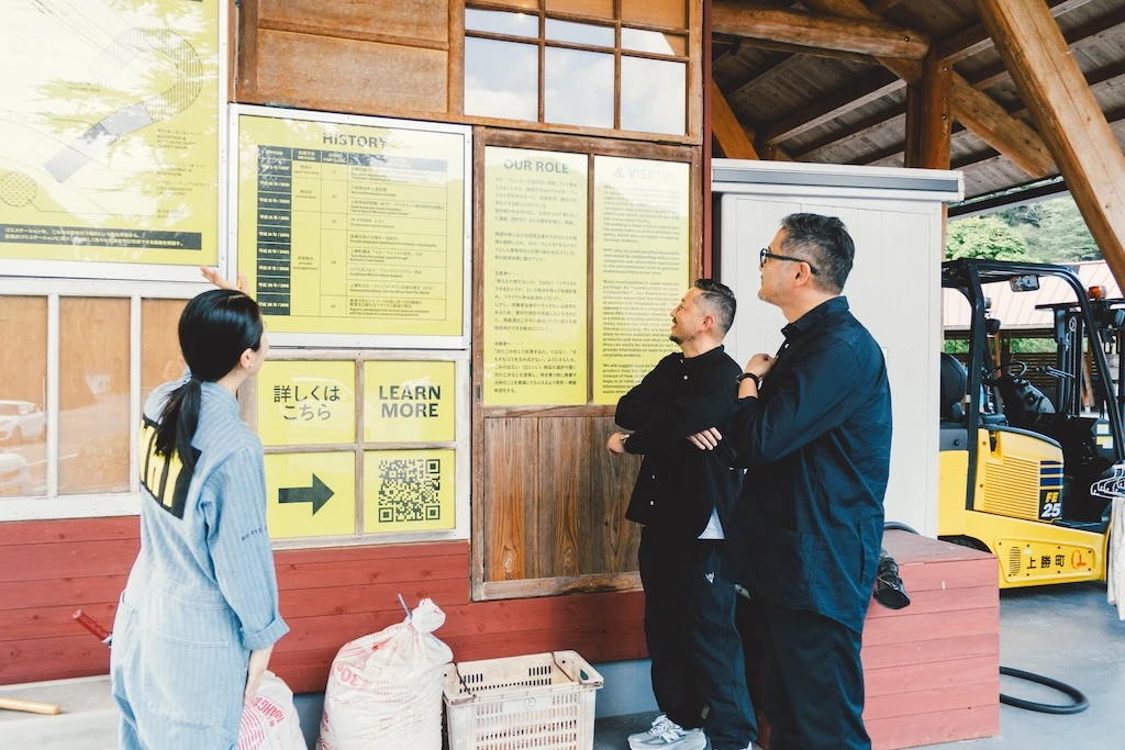 Ms. Irobe and Mr. Watanabe reading the information posted on the wall of the trash station; Ms. Otsuka and others are seen explaining something to them.