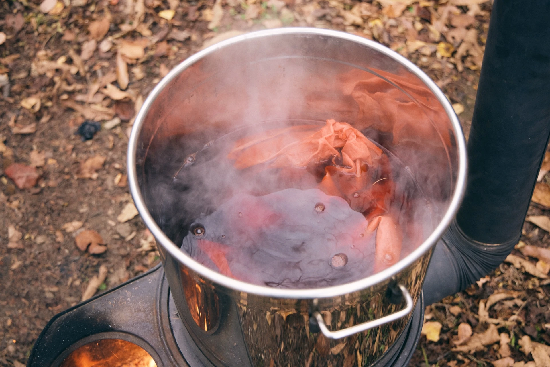 A photograph of a silver pot containing water and cloth with dye in it. Steam is coming out of the pot, indicating that it is heated by a wood stove.