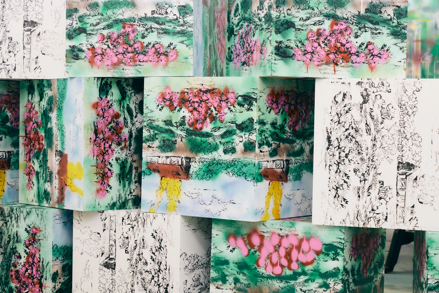 A close-up photo of a randomly stacked pile of cubes. All sides are silkscreened with images of houses in nature. Some cubes are sprayed or painted, while others feature uncolored line drawings.