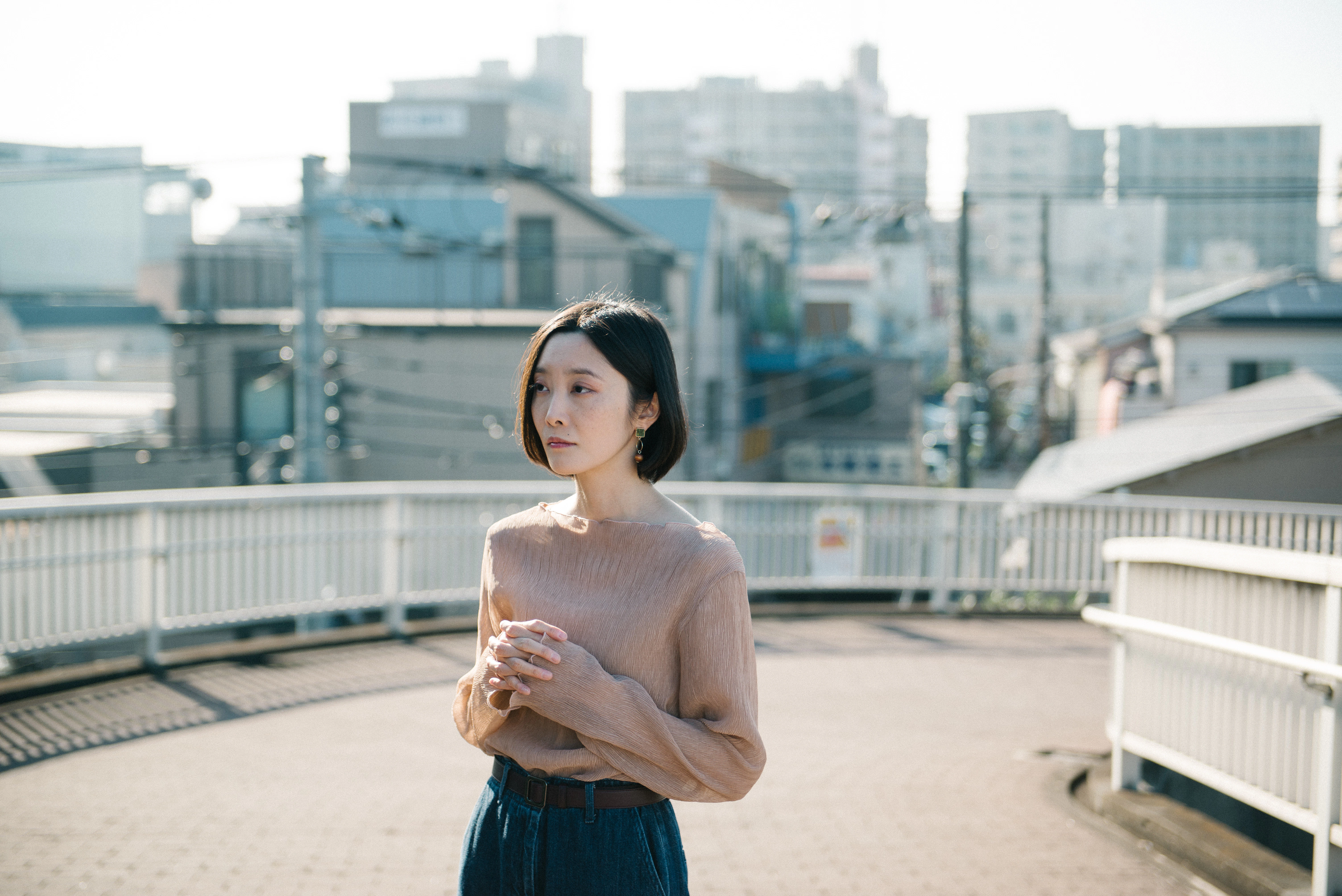 ポートレイト。ベージュのブラウスに黒いパンツ。歩道橋の中央で、胸の前で手を組み立っている。