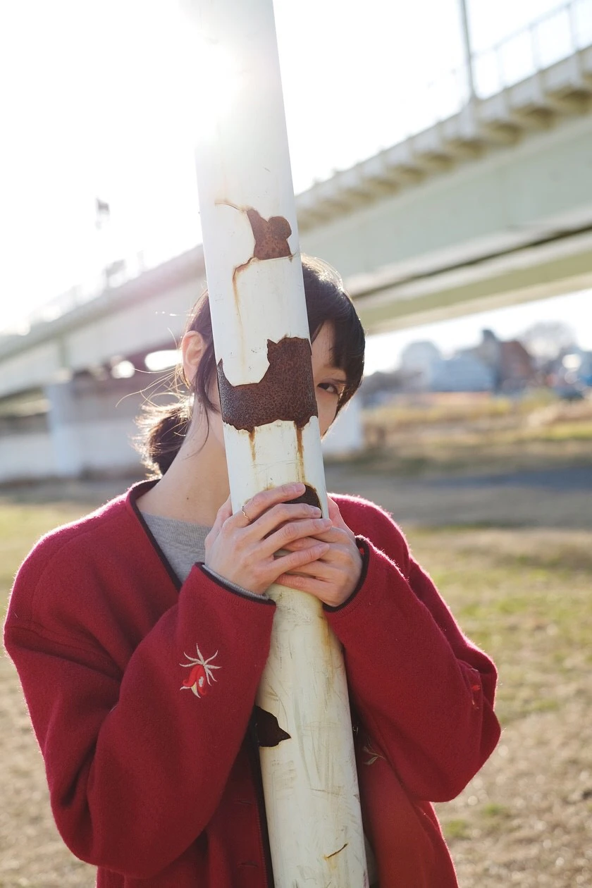 鶴田理紗の宣材写真。背景には河川敷の芝生、川をまたいで電車の線路がある。手前に錆びついた白いポールがあり、鶴田が後ろからポールを両手で掴んでいる。ポールで顔の半分が隠れている。花の刺繍があしらわれた赤いコートを着用している。