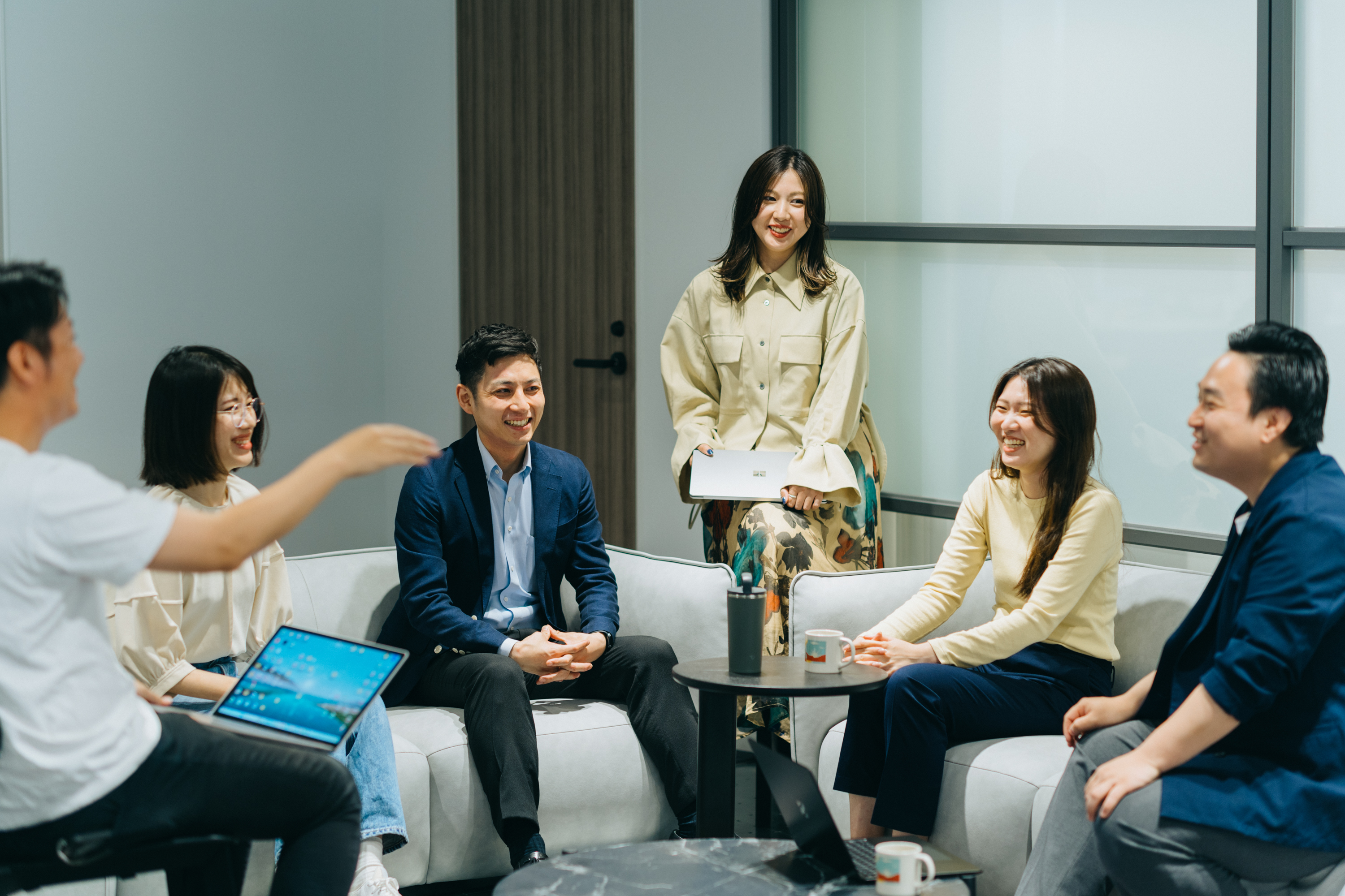 丸い机を囲むソファー席に座って談笑する6人の社員の写真。