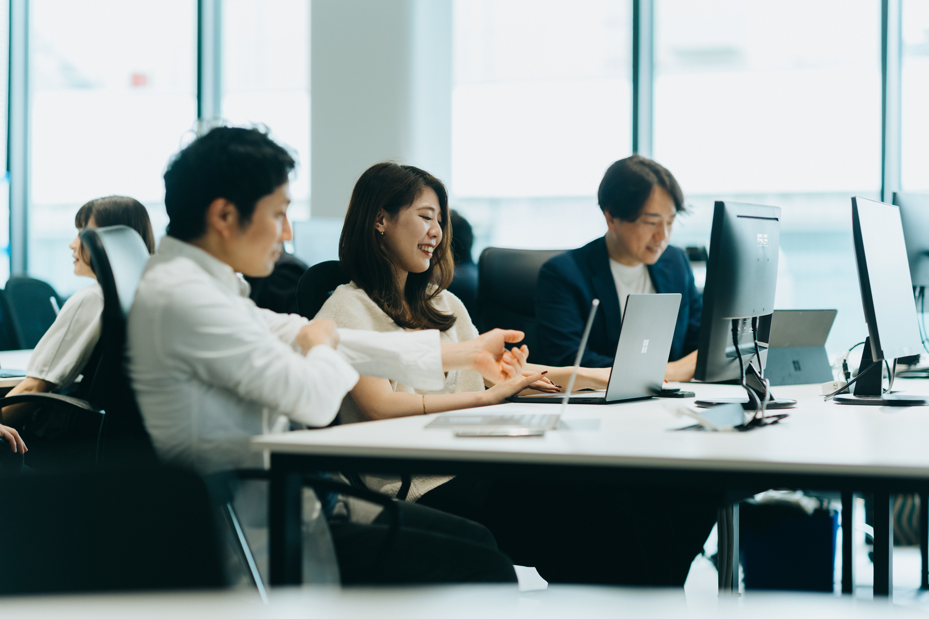 オフィス内の長机に3名の社員が並んで座り話し合いながら作業をしている写真。